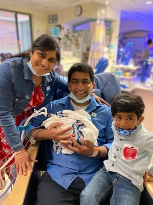 Mother standing, father seated holding infant bundled in blanket and young son sitting next to father all posing for a picture in a blurred hospital area in the background.