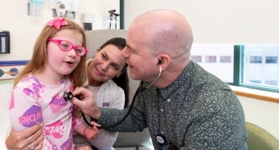 A clinician uses a stethoscope on a young girl wearing pink glasses and sitting in an adult woman's lap in a medical exam room.