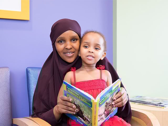 Mother wearing a dark head scarf with her daughter on her lap. The daughter is holding a book open.