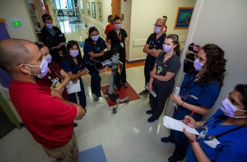 A group of several masked clinicians stands talking in a hospital hallway.