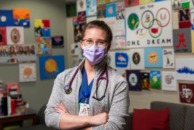 A young, masked woman with a stethoscope around her neck stands with her arms crossed, smiling