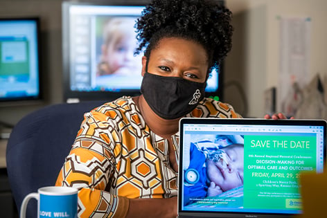 Telisa Hassen wearing a black Children's Mercy face mask and smiling with her eyes. She is sitting behind a computer monitor that has a Children's Mercy "save the date" graphic that she designed.
