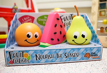 Fruit Friends toys: an orange, a slice of watermelon and a pear. The orange and the pear have eyes and a smiling mouth. Packaging reads, “Fruit Friends, Nourish the Senses, Fat Brain Toy Co.”