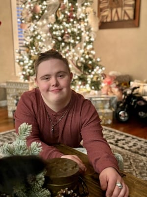 A young adult man wearing a maroon shirt sits on the floor in front of a Christmas tree.