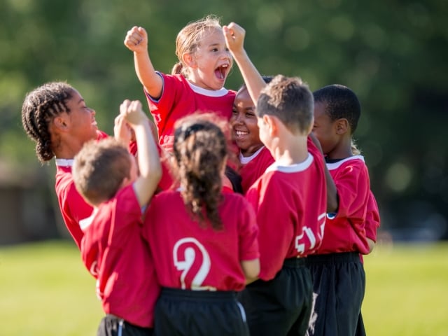 A group of pre-teen girls and boys in red soccer uniforms celebrate together by lifting up a girl at the center of their circle.