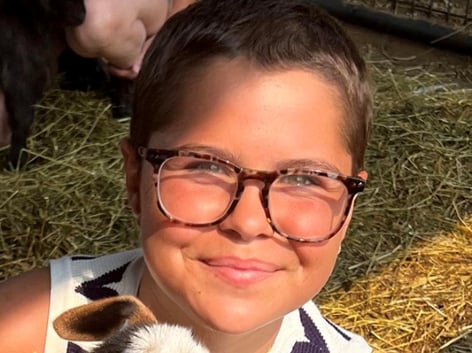 A teenage girl with short brown hair and glasses holds an animal with hay in the background.