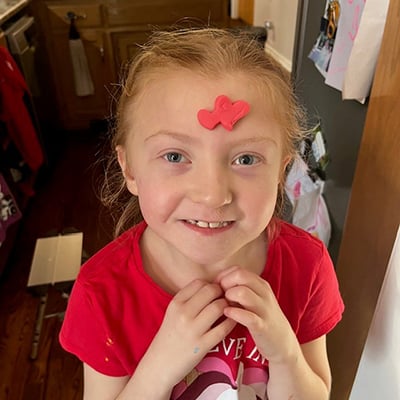 A young girl with red hair pulled into a ponytail poses with a red piece of clay stuck to her forehead