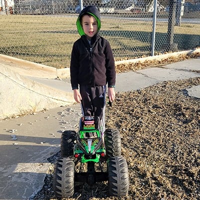 A young boy standing on a sidewalk beside a large green remote-controlled monster truck, wearing a dark jacket and hood.