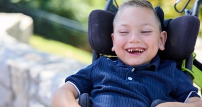 Lucas Dahmer sits in a wheelchair laughing. He is outside. He is wearing a navy blue shirt with buttons at the top and a collar.