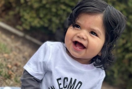 A smiling baby with curly black hair wears an ECMO t-shirt.
