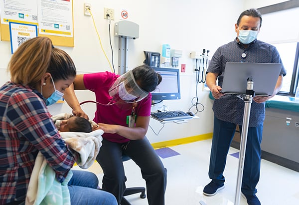 A man who is a language coach stands in front of a stand which holds his laptop computer, while a female clinical staff person listens to the heartbeat of an infant on a baby’s back with her stethoscope, while mom holds the baby in her arms wrapped in a blanket