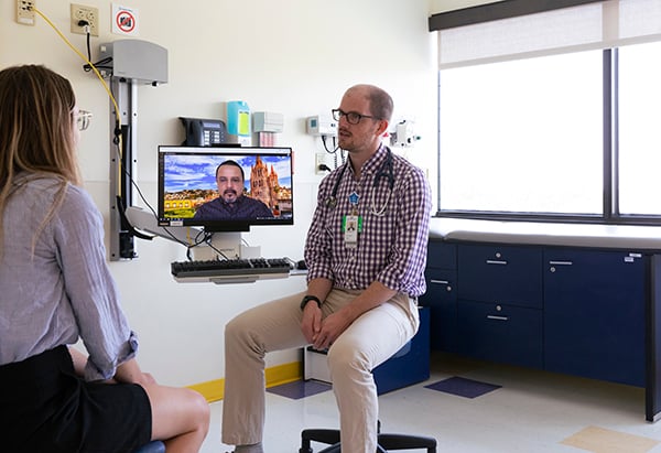 a physician sits on a stool in a clinic’s examination room speaking to a female patient who appears to be a teen, while a Cultural Language Coaching Program male staff member’s face appears on a computer screen virtually to assist the physician with language coaching.
