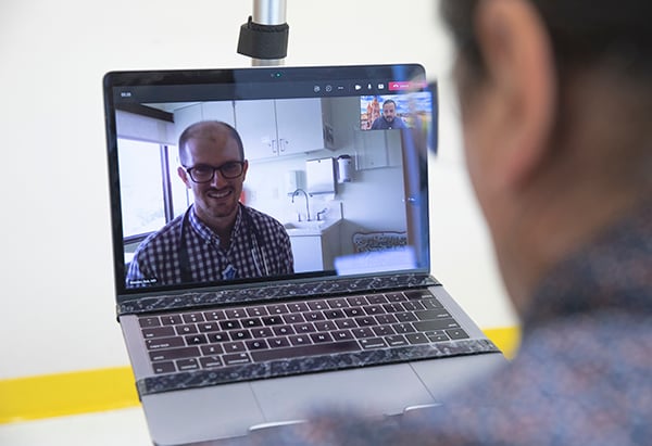 a CM physician’s face is on a laptop computer screen smiling and a Cultural Language Coaching Program male staff member is also on the laptop computer screen in a small box in the top right-hand corner.  Both are virtually meeting with a man who is holding the laptop computer, with only the back of his head shown.