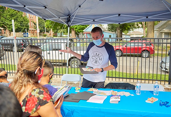 a male in a mostly white t-shirt w/navy blue mid-length sleeves with a CM logo on the front, is holding a black folder and colorful kiddie stickers in one hand.  There are 5 people lined up at his table where he is handing out pamphlets and information on healthcare