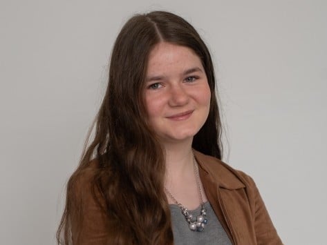 A teenage girl with wavy brown hair is wearing a gray shirt and a brown jacket.