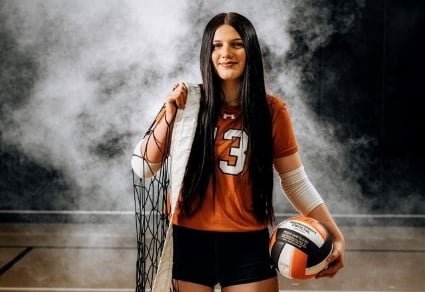 A teenager wears an orange and black volleyball uniform and holds a net and ball. She is standing on a volleyball court and there is dramatic smoke behind her.