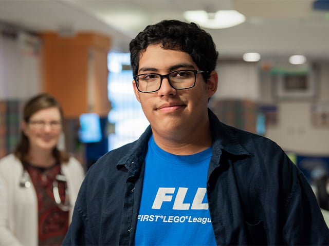 Teen boy patient grinning with a Children's Mercy provider blurred in the background. They are inside Children's Mercy Adel Hall hospital.