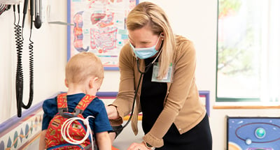 Dr. Sarah Edwards wearing a face mask and using a stethoscope to listen to the the lungs of a patient wearing a backpack with medical equipment inside