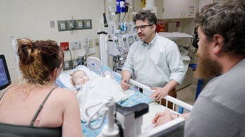 A child lies in a hospital bed covered with a blanket, connected to medical devices. Three adults are present—one standing beside the bed speaking, and two on either side, listening. The room contains medical equipment, monitors, cabinets, and supplies, creating a clinical setting.