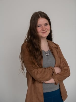 A teenage girl with long, wavy hair stands against a plain background. She is wearing a brown jacket over a gray shirt, with arms crossed in front of the chest, and a necklace with multiple pendants.