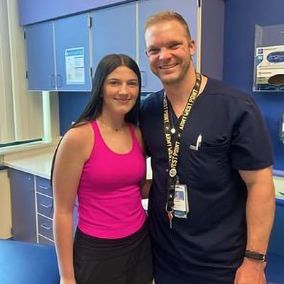 A teen with long hair and a pink sports top stands next to a doctor in navy blue scrubs and a hospital employee lanyard and ID card.