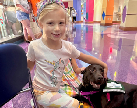 Children's Mercy patient, Charley. She is smiling and sitting on the floor at Children's Mercy Hospital with Facility Dog, Litta.