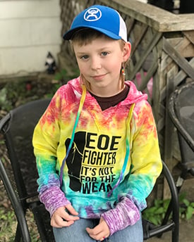 Austin Hasenohr sitting in a chair outside, grinning, and wearing a tie dyed sweatshirt that reads, "EOE fighter, it's not for the weary."