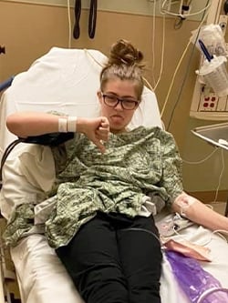 Taylor Stewart laying on a hospital bed with a gown on sticking out her tongue and giving a "thumbs down."