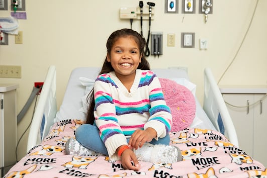 A smiling girl with long brown hair sits cross-legged on a hospital bed.