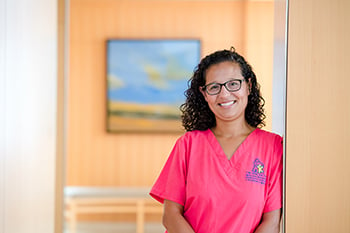 Amy Cobos smiling and leaning against an open door frame at Children's Mercy hospital. She is wearing a hot pink Children's Mercy scrub top with the Children's Mercy logo.