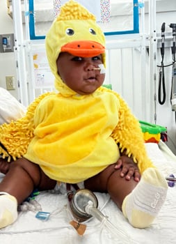 A Black baby is dressed in a yellow chicken costume for Halloween. He is sitting in a hospital bed with a tube coming from his nose and other tubes and medical equipment around him.
