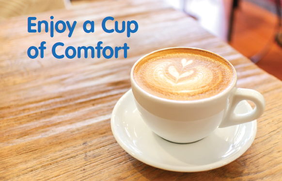 White cup with saucer on a table. The cup is filled with coffee with foam on top in the shape of two hearts. Words read: enjoy a cup of comfort.