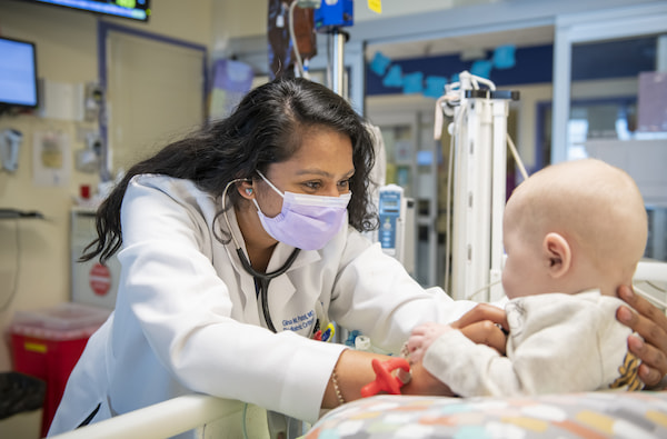 A masked, female physician examines a young toddler in a hospital bed.