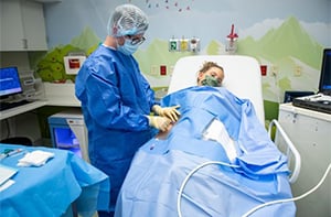 A A Children's Mercy nurse wearing a blue gown, gloves, and a hat  and a young patient wearing a face mask and covered with a blue sheet. The nurse is giving the patient numbing medicine through the arm.
