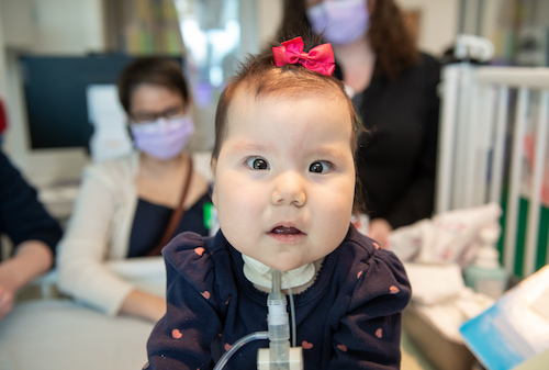 A young girl with a tracheostomy tube and a pink bow in her hair looks directly at the camera.
