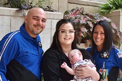 Willow Noud held by her mother with Transport team members on each side, posing in front of the annex building in the Adele Hall Campus circle drive.