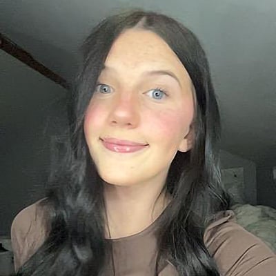 A teenager smiles at the camera. Her hair is long, dark and curled and her eyes are blue. She’s wearing a brown shirt.