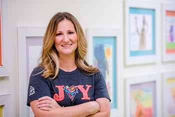 Amy Findlay smiling and wearing a Children's Mercy shirt that reads, "JOY" with the "o" being a heart shape. She is  at Children's Mercy and standing in front of multiple artwork done by CM patients.