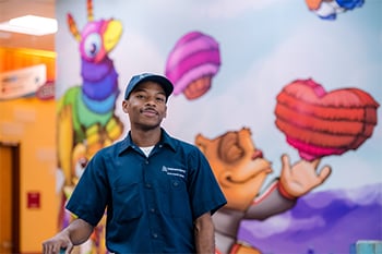 Nick Suber is smiling and wearing a blue Children's Mercy uniform (shirt and cap). He is standing in Children's Mercy hospital with a colorful mural of cartoon characters behind him on the wall.