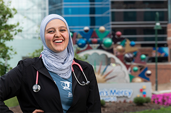 Zainab Al-Janabi is smiling and standing outside in front of a Children's Mercy hospital sign. She is wearing a hijab and stethoscope around her neck.