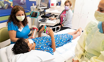 A boy in a hospital gown laying on an examination bed at Children's Mercy. His legs are straight on the bed. Three Children's Mercy providers are on either side of the bed.