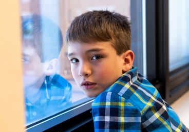 A young boy in a plaid shirt looks at the camera and is reflected in a nearby window
