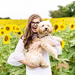Taylor Stewart standing with a big smile in a field of sunflowers while holding a dog. She is wearing a white dress and eyeglasses.