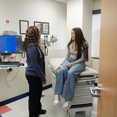 Two people are in a doctor’s office exam room. One is standing and wearing a blue hoodie and black pants. She has wavy brown hair with highlights and is talking to the patient. The patient is a teen girl with long brown hair with highlights. She is seated on the exam table and wearing a gray shirt and jeans.