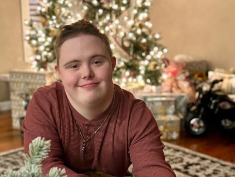 A young man with brown hair and a maroon shirt sits on the floor in front of a Christmas tree.
