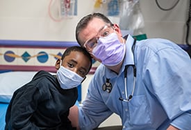 Chase Jackman and Dr. Joel Thompson with their arm around each other and heads touching while looking at the camera. They are wearing face masks and inside a Children's Mercy patient room.