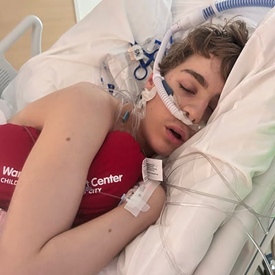 A teenage boy lies in a hospital bed with medical tubing visible and holds a heart shaped pillow against his chest.