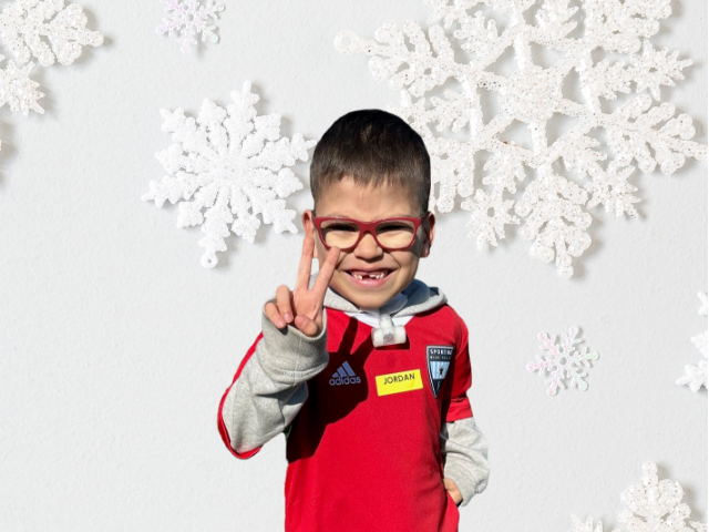 A young boy in a red shirt and red glasses, with a tracheostomy tube, holds up two fingers. He is standing in front of a white background with snowflakes.