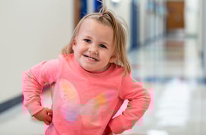 A young girl with a ponytail on top of her head and a pink shirt stands with her hands on her hips.