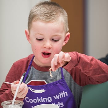Boy wearing a purple apron that reads “Cooking with Keto Kids.” He is holding pasta in one hand and a spoon in his other.